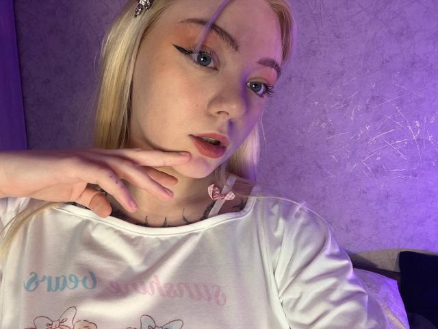 EnchantingEyes with blonde hair, blue eyes, and thick black eyeliner, touches her chin with her right hand, wearing a white shirt, under purple lighting.