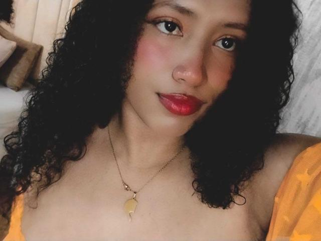 KarinaLovey has curly black hair, red lipstick, and a gold necklace with a leaf pendant. She's topless with orange patterned sleeves, showing her tits. Her nose is pierced.