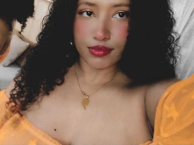 KarinaLovey with curly black hair, red lipstick, and a gold heart necklace, wearing an orange off-shoulder top, showing her bare tits and a small black mole on her chest.