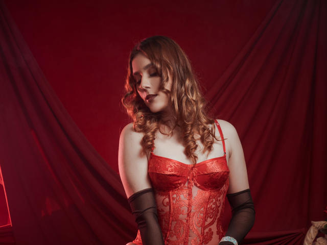 GoddessBar avec des cheveux roux bouclés, portant un corset en dentelle rouge et des gants noirs, les yeux fermés, les bras baissés, fond rouge, montrant ses seins, avec une allure sexy et confiante..