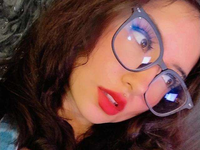SusanMonroy has red lips, a nose piercing, and wears thick glasses with blue eyeliner. Her brown hair is wavy, and she's looking up with a slightly open mouth.