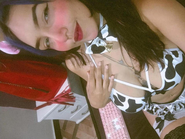 SophieWolf in un bikini a pois bianchi e neri, con le guance rosse, capelli neri con riflessi viola, che mostra il seno con una mano vicino al petto, indossando un collare con campanellini..