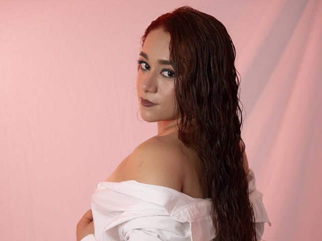 ValeryHilton with wet brown hair, looking over shoulder, wearing an off-shoulder white shirt, showing bare shoulder and subtle cleavage. Pinkish background. She has dark lipstick and a necklace.