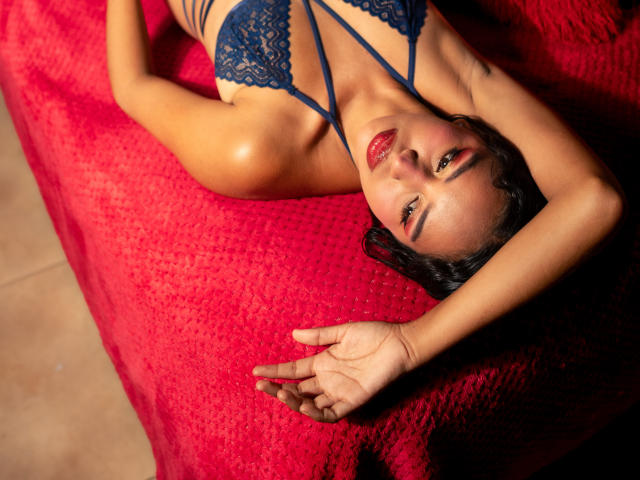 JeimyCampos, lying on a red textured bed, wearing a blue lace bra, arms above head, red lipstick, dark hair, and a sexy, relaxed expression, with visible cleavage.