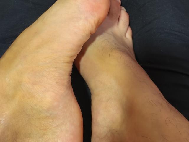 OrianaLaFrancaise's toes are intertwined with her ankle, showing slightly hairy skin on the bottom. Her foot is slightly pink, with visible skin texture. Gray bottom.