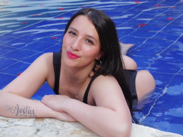 SamyNoa is in a pool, lying on her stomach with black one-piece, showing her ass, and has pink lipstick, black hair, and a tattoo on her left arm.