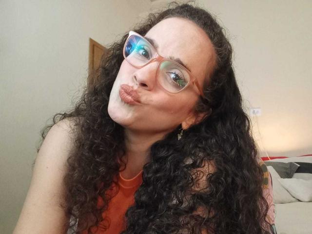 AmyFlores, met krullend zwart haar, draagt een rode bril en een rode top, ze frumelt haar lippen in een selfie en laat haar bleke huid en kleine, schattige neus zien..