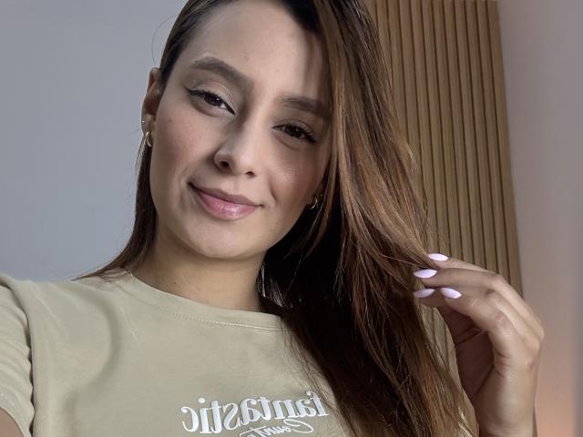 MyaSpecter has light brown skin, long brown hair, and a small smile. She's wearing a beige shirt, holding her hair with pink nails, and has a cute, confident look.