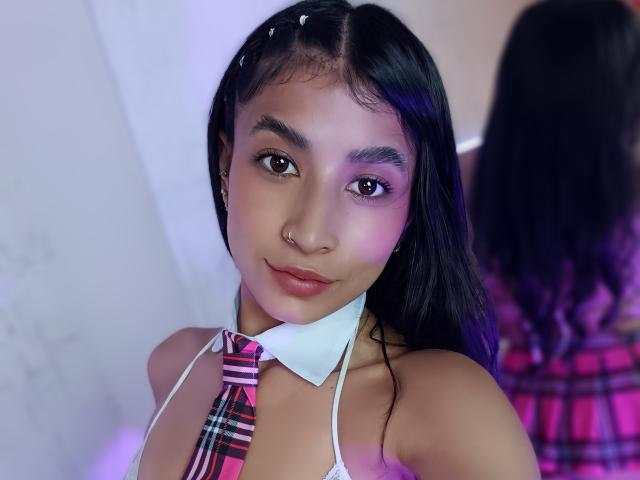 NaiaSanders with long black hair, wearing a plaid necktie and white halter top, showing her small nose ring and subtle smile, reflected in a mirror.