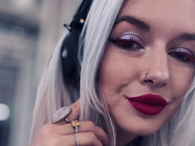 EnaMore has pale skin, silver hair, red lipstick, purple eyeshadow, and a nose ring. She's wearing black headphones, rings, and a yellow ring with a gem. Smiling subtly.
