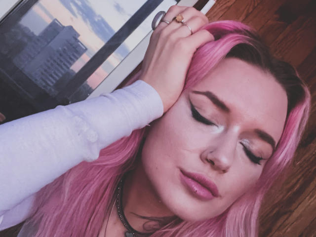 EnaMore with pink hair, closed eyes, and thick black eyeliner, holding her head with a ringed hand, wearing a white long-sleeve shirt, looking pensive.