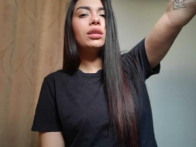 AmaiaSalvatoree with long black hair, pouty lips, black tee, raising arm, tattoo on upper arm, light brown skin, standing against a wooden and white background.