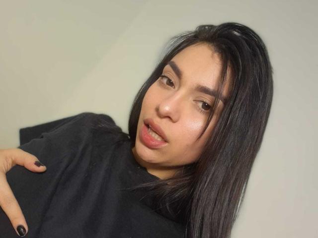 AmaiaSalvatoree with black hair, light skin, brown eyes, and black nails, wearing a black shirt, looking slightly to the side with a playful, open-mouthed expression.