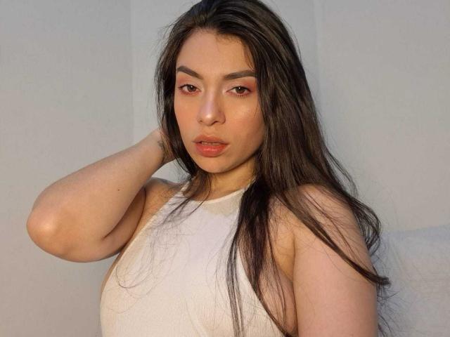 AmaiaSalvatoree has long dark hair, pinkish-red eyes, and is wearing a white sleeveless top, with one hand behind her head, showing her soft, slightly parted lips.