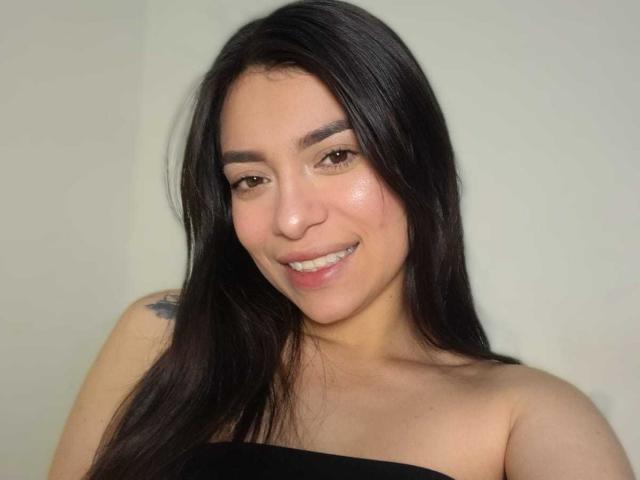 AmaiaSalvatoree with long black hair, smiling, bare shoulders, black strapless top, light skin, small tattoo on left shoulder, brown eyes, subtle makeup, slightly shiny skin.