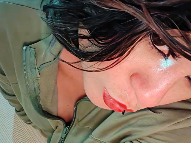 CassieJones has wet black hair, shiny skin, red lipstick, and is wearing a green jacket with a zipper, looking up with a slightly parted mouth. Boobs partially visible.