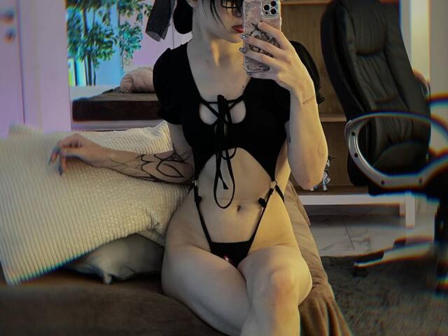 SimaWendy with pale skin, black lace-up top, small tits, black thong, tattoo on arm, taking mirror selfie, sitting on brown couch, holding phone with pink case.