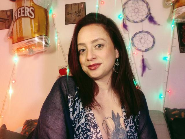 CatalinaMonyque, con lápiz labial rojo, ojos marrones y cabello castaño oscuro, vestida con una blusa azul estampada, mostrando un tatuaje debajo de los senos, sonriendo a la cámara..