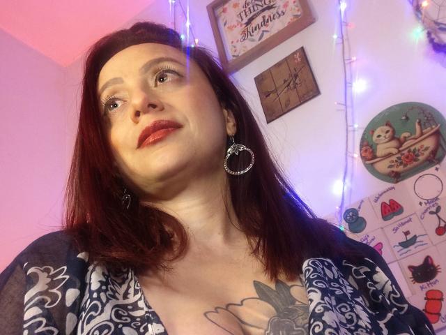 CatalinaMonyque, con lápiz labial rojo, cabello oscuro y un tatuaje en el pecho, vestida con una blusa azul estampada y grandes aros, mirando hacia arriba con una ligera sonrisa..