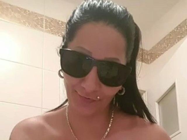 Brighy has dark hair, black sunglasses, a nose piercing, and a gold necklace, smiling with bare shoulders, wearing a gold hoop earring, and a subtle lip piercing.