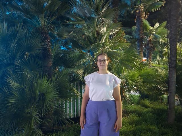 BellGabi stands in front of palm trees, wearing a white short-sleeve shirt and purple pants, with glasses and a calm expression, in a sunlit, green garden.