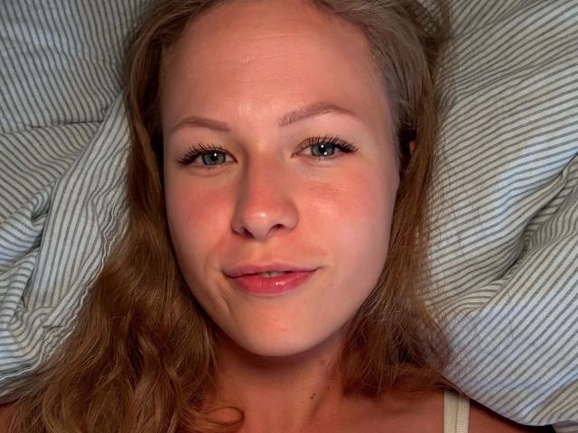JuliaMey-hot has blue eyes, wavy brown hair, fair skin, and a slight smile. She's lying on a striped pillow with her head slightly tilted to the right.