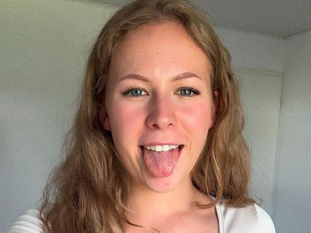 JuliaMey-hot has wavy brown hair, blue eyes, and a playful expression with tongue out. She's wearing a white top, showing her pink-tinted nose and subtle freckles.