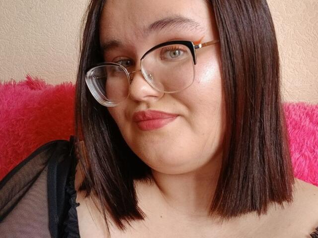 JessicaJoye has shiny skin, dark brown bob, and wears glasses with black frames. She's smiling with pink lipstick, black mesh top, and pink furry pillow behind her.