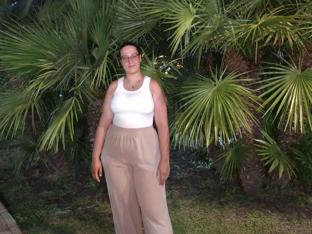 BellGabi has medium tits, a white tank top, beige pants, and glasses, standing in front of green palm trees at night. Her ass is average-sized, and she's smiling.