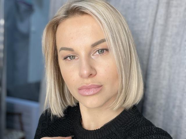 BelleLisaG with blonde bob, green eyes, and pinkish lips, wearing a black knit sweater, looking straight at the camera. She's got a cute, sexy face.