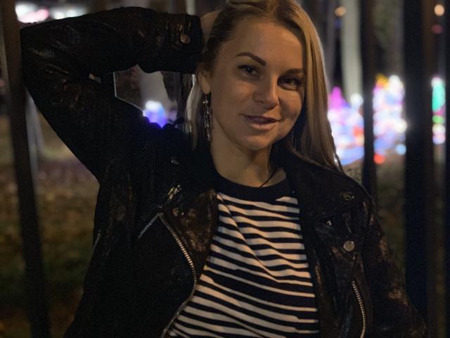 BelleLisaG with blonde hair, black lace jacket, striped shirt, and purple lipstick, hand in hair, smiling, bokeh lights in background, night, dark setting, earrings visible.