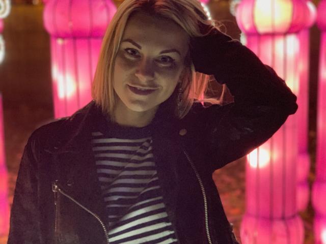 BelleLisaG with blonde hair, wearing a black jacket and striped shirt, hand in hair, smiling at camera, pink lanterns in background, soft pink and purple lighting.