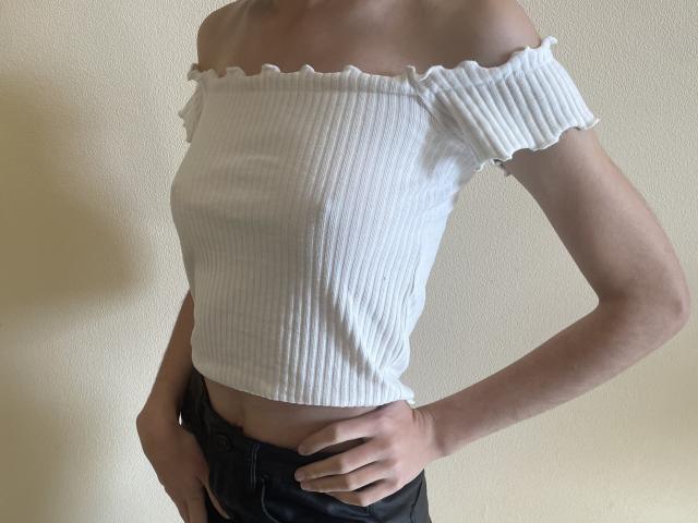 Millinia wearing a white, ribbed off-shoulder top, showing small tits, with black pants, hand on hip, light skin, and a slim, sexy, feminine build.
