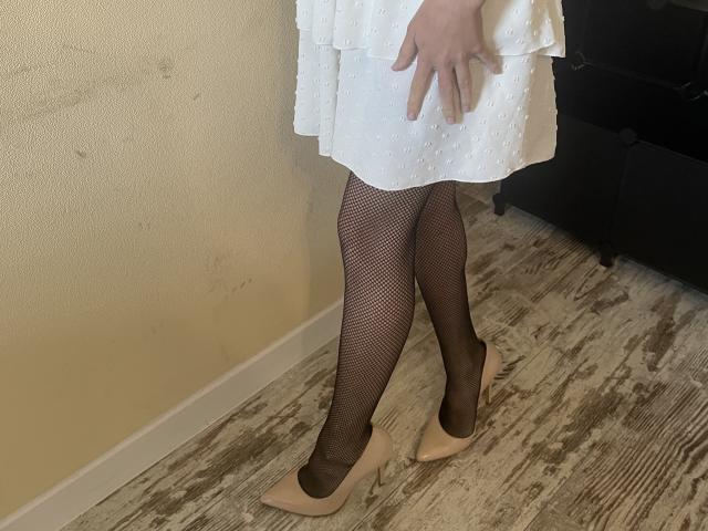 Millinia's legs in fishnet stockings, white dress with dots, and nude heels. Her right hand rests on her thigh. Sheer black fishnets show her skin tone.