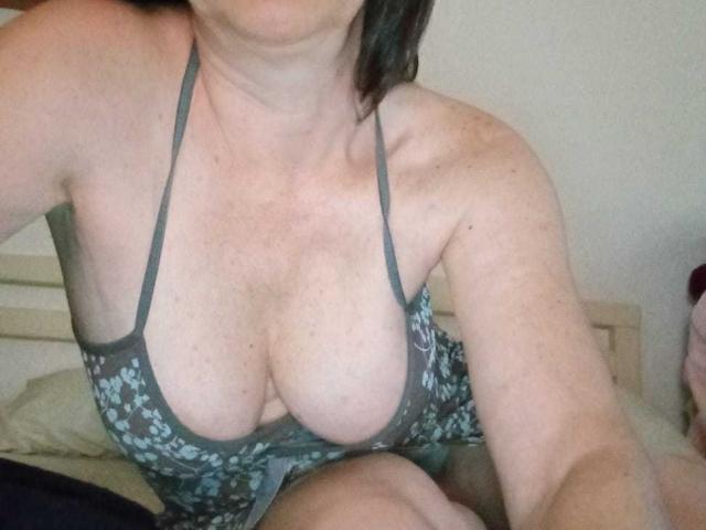 MilfLhara has large, pale, freckled tits in a grey floral halter dress, showing significant cleavage, with her arm and head partially in the frame, on a bed.