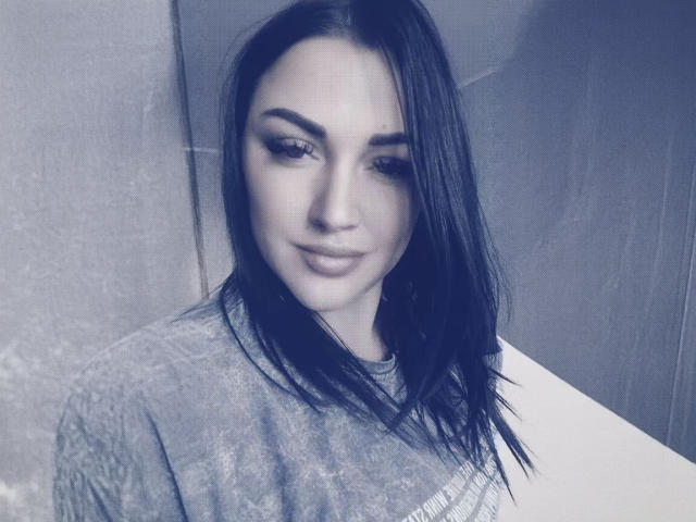 XCrazyBabeX with dark, straight hair, wearing a grey t-shirt, has a slight smile, and is looking downward with subtle makeup highlighting her eyebrows and eyeliner.