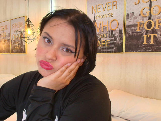Blueeskyy with black hair, pink lipstick, and black shirt, pouting with hand on cheek, brown eyes, wearing makeup, in a cozy room with hanging lamp and city posters.