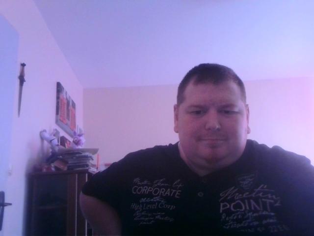 LoveDoux is a chubby, short-haired dude with a slight smile, wearing a black shirt, standing in a dimly lit room with purple and pink lighting.