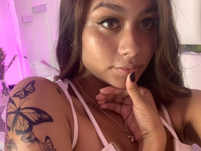 KinkyCuteLady with brown hair, freckles, and black nail polish, touching her chin, wearing a pink top, showing a butterfly tattoo on her left shoulder, and a necklace.