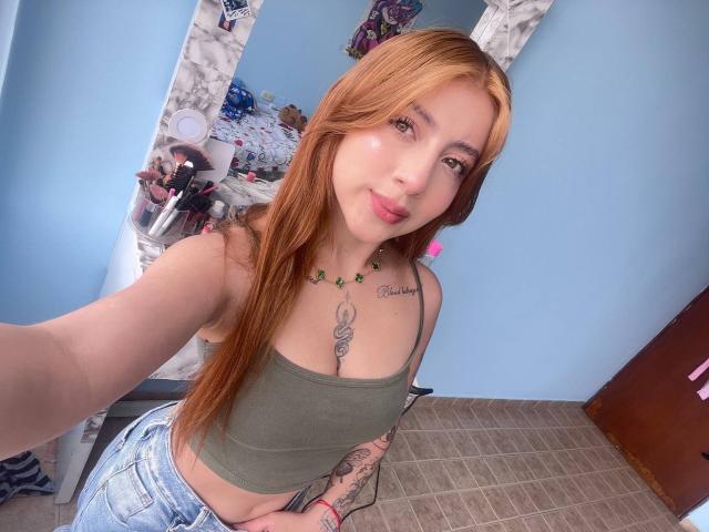 CharlotteAristizabal, with long red hair, green crop top, and blue jeans, showing a tattooed chest and small tits, smiling in a mirror selfie. Her hand rests on her hip.