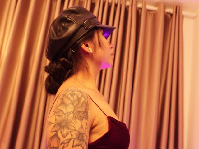 MilaDor con un gorro de cuero negro, luz púrpura en su rostro, un brazo tatuado y un escote pronunciado en un vestido rojo, de pie de lado, mostrando sus senos..