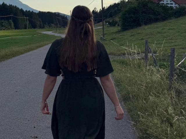 Bailey-hot with long brown hair in a braid, wearing a black dress, standing with arms slightly out, on a gravel path, facing away, in a green, grassy, rural area.
