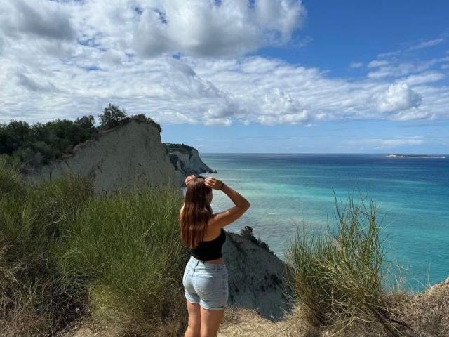 Bailey-hot with long brown hair, black tank top, and blue shorts stands on a cliff, hands on head, looking at a blue ocean with white clouds.