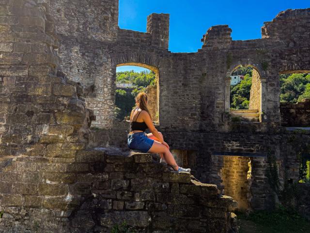 Bailey-hot with a tan ass, wearing a black top and denim shorts, sitting on stone ruins with her back to the camera, under a bright blue sky.