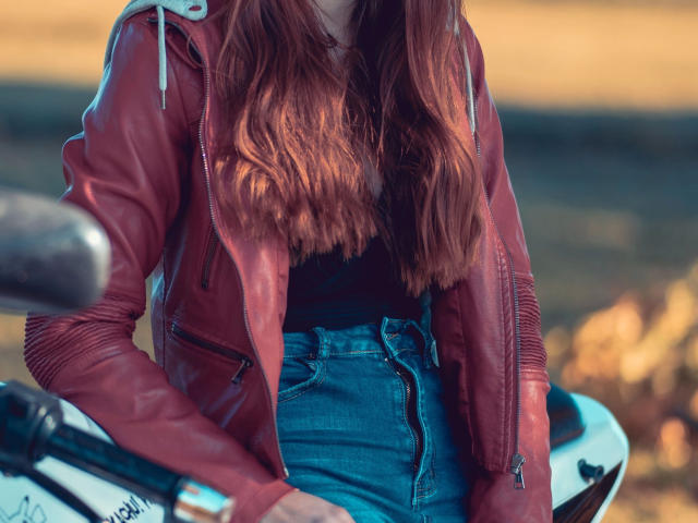 Bailey-hot with red leather jacket, blue jeans, and long brown hair, sitting on a white motorcycle, hands on seat. Her ass is partially visible. Soft sunset light.