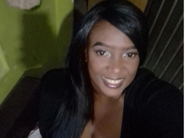 SofiaSims with dark, shiny hair, smiling with shiny, glossy lips, wearing a black top, showing cleavage, brown eyes, and smooth skin, in a dimly lit setting.