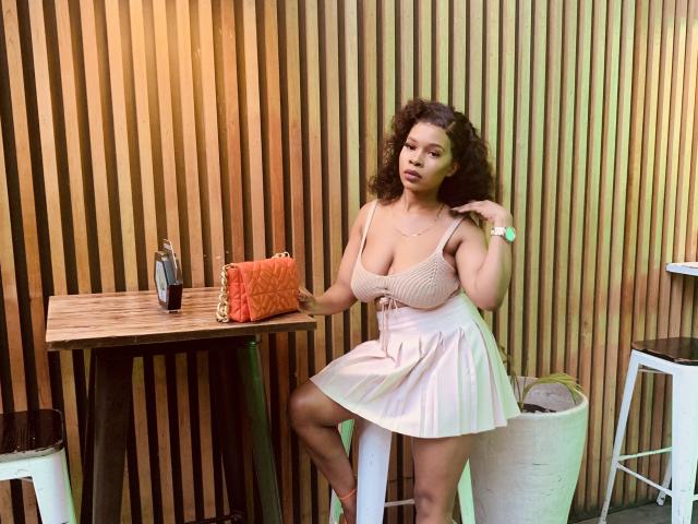 DorothyKatty with curly hair, big boobs in a pink dress, sitting on a stool, hand on her shoulder, wooden wall, orange purse, bottle, and white stool in background.