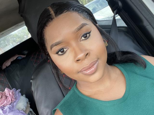 DorothyKatty's face, with glossy pink lips, dark straight hair, green top, and winged eyeliner, sitting in a car, showing off her cute ass and small, shiny nose.
