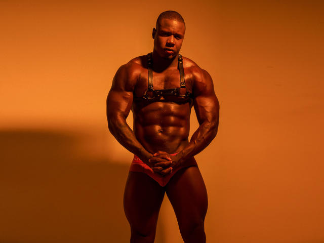 SexyBlackym is a muscular black man with a shaved head, wearing a black leather harness and red underwear, standing with hands in front, under warm orange lighting.