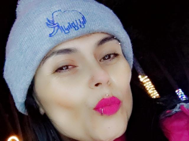 CharlotteCrofort with pale skin, pink lipstick, and a silver nose ring, pouting with black eyeliner, wearing a blue knit beanie, and a pink jacket, in a dark setting.
